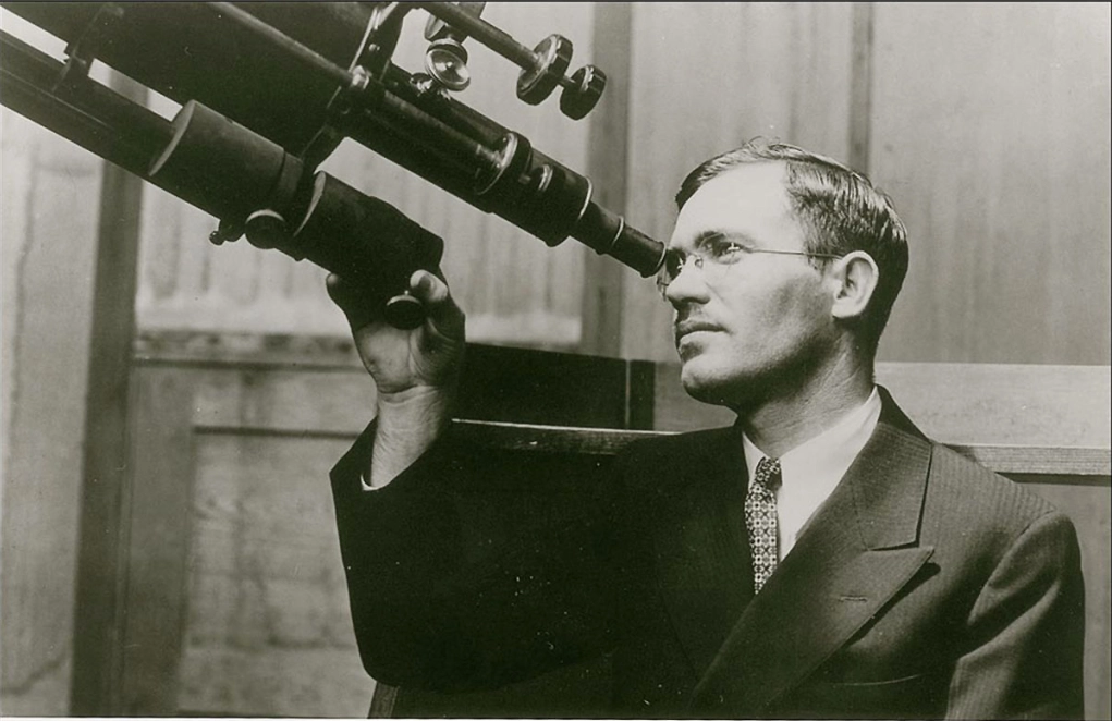 A black and white photo of Clyde Tombaugh looking through a telescope.