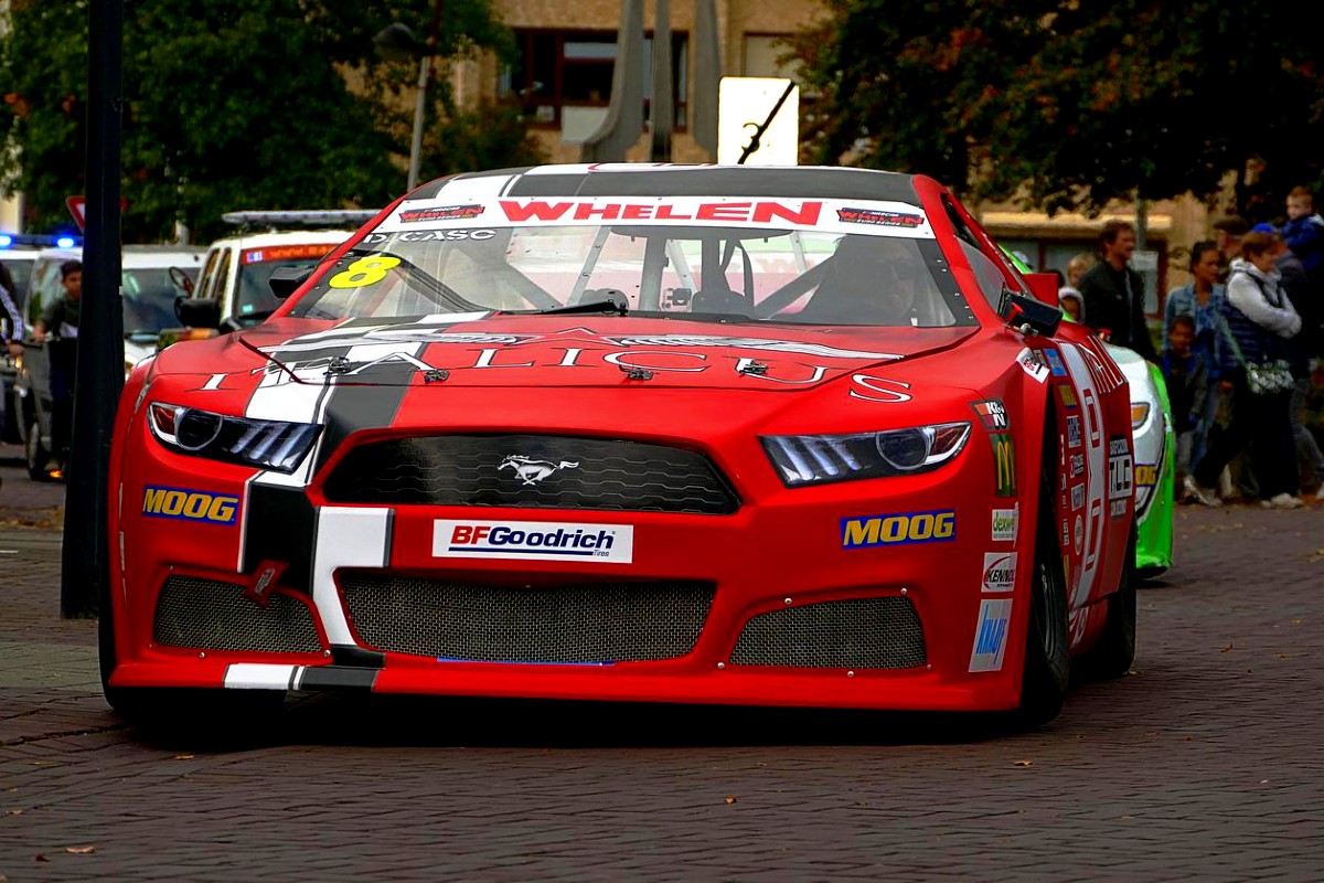 A red NASCAR-branded Ford Mustang driving past a group of bystanders